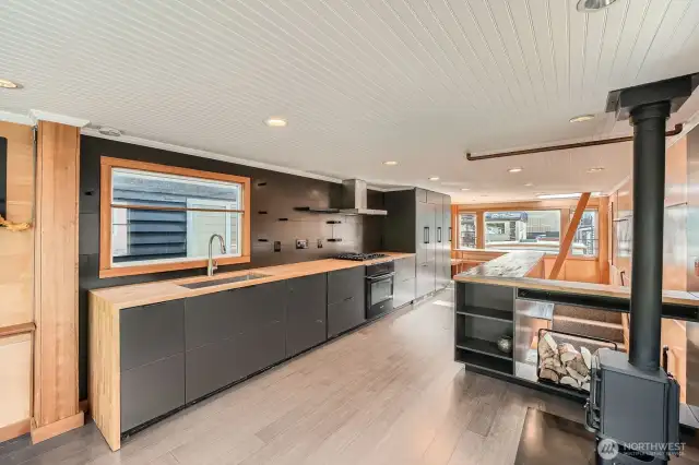 This sleek, modern kitchen stuns with clean lines, hidden appliances, and warm butcher block counters. Matte black cabinetry blends seamlessly with integrated storage, while the gas range and minimalist open shelving complete the contemporary design. A cozy wood stove nearby adds charm and warmth—perfect for life on the lake.