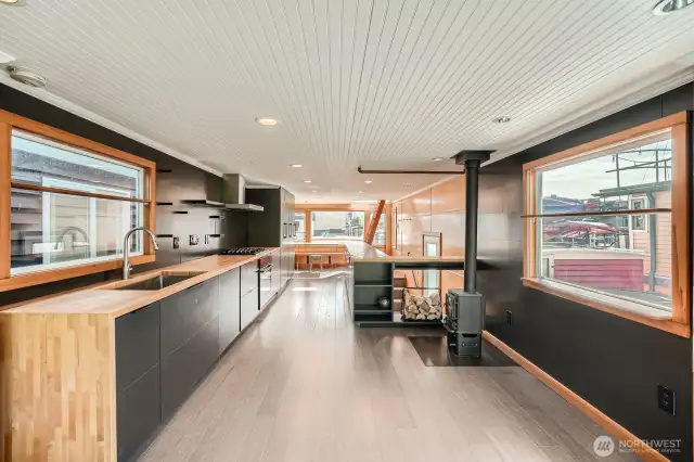 This photo highlights the cozy wood-burning stove set against a sleek matte black wall, adding warmth and character to the modern space. A butcher block island with open shelving offers both function and style, while the unique peekaboo window reveals the stairwell below—an inviting architectural detail that enhances light and flow.