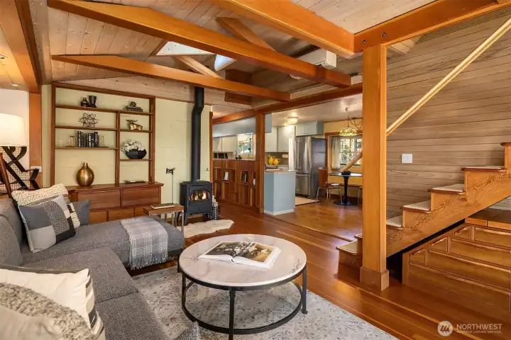 The living room looking toward the kitchen — wood stove, built-in shelving, vaulted beams, and a sectional sofa that invites you to settle in and stay a while.