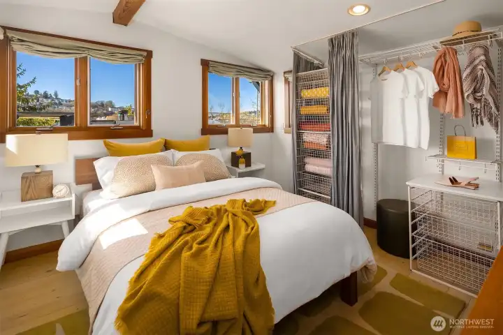 The upper-level primary bedroom is bright and airy, with dual corner windows, warm wood accents, and an open wardrobe system that makes getting ready a pleasure.