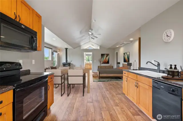 This view highlights the spaciousness of the open-concept living area, featuring vaulted ceilings, a ceiling fan, and a mini-split system. It includes a dining space, full kitchen, and ample room for your creative design ideas. The layout also offers an attached bath, two closets (one walk-in), and additional built-in wall storage for maximum convenience. The free-standing wood stove provides cozy warmth on cool nights.