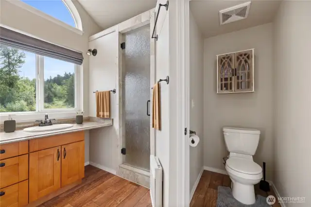 The attached bath in the open-concept living space features a luxurious 8.5-foot walk-in shower with dual rain shower heads and a built-in bench. Large windows flood the space with natural light, complemented by a custom window shade for privacy. A separate toilet room with its own door adds convenience, while the vanity offers ample storage. Additional features include a spacious closet, radiant wall heat, and durable LVP flooring for both comfort and style.