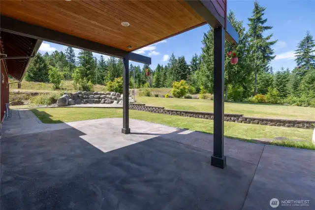 This view highlights the upper-level balcony, which provides a shaded patio area below—perfect for relaxing on warm days. A block wall elegantly separates the patios from the expansive, landscaped yard, which features a sprinkler system. Additionally, a separate cobblestone patio bordered by a striking large rock wall offers an ideal space for a fire pit and outdoor gatherings.