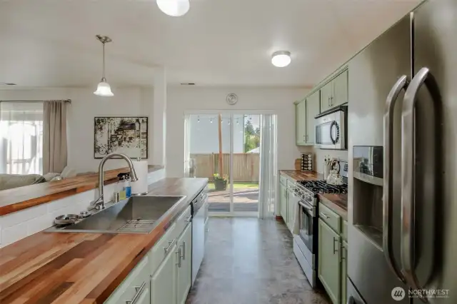 This stunning kitchen showcases custom butcher block countertops, under-cabinet lighting, stainless steel appliances, a large sink, and a breakfast bar for guests—plus easy access to the back deck through the sliding glass door.