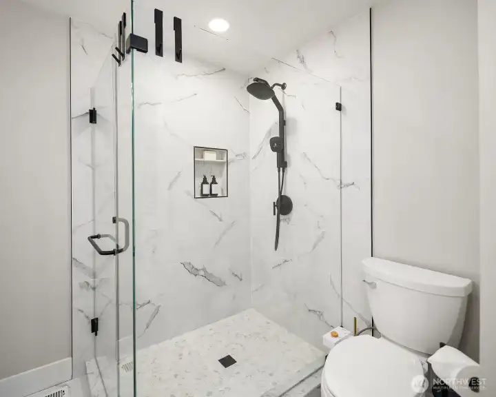 A stunning walk-in shower with full granite slabs on the walls, a niche for soap, dual shower head with a handy wand, tile flooring and glass doors.
