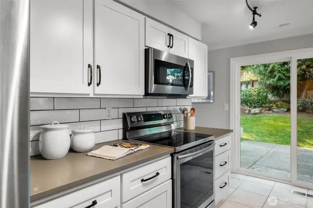 Bright white cabinets and backsplash complement the spacious design, while a large sliding glass door invites the outdoors in, bringing in all the natural greenery.