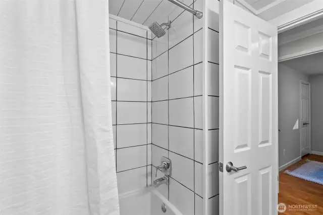 Light and bright, this bathroom features sleek white tiles, stylish black fixtures, a durable vinyl plank floor, skylights, a shiplap ceiling, and a large frosted window ensuring privacy without sacrificing natural light.