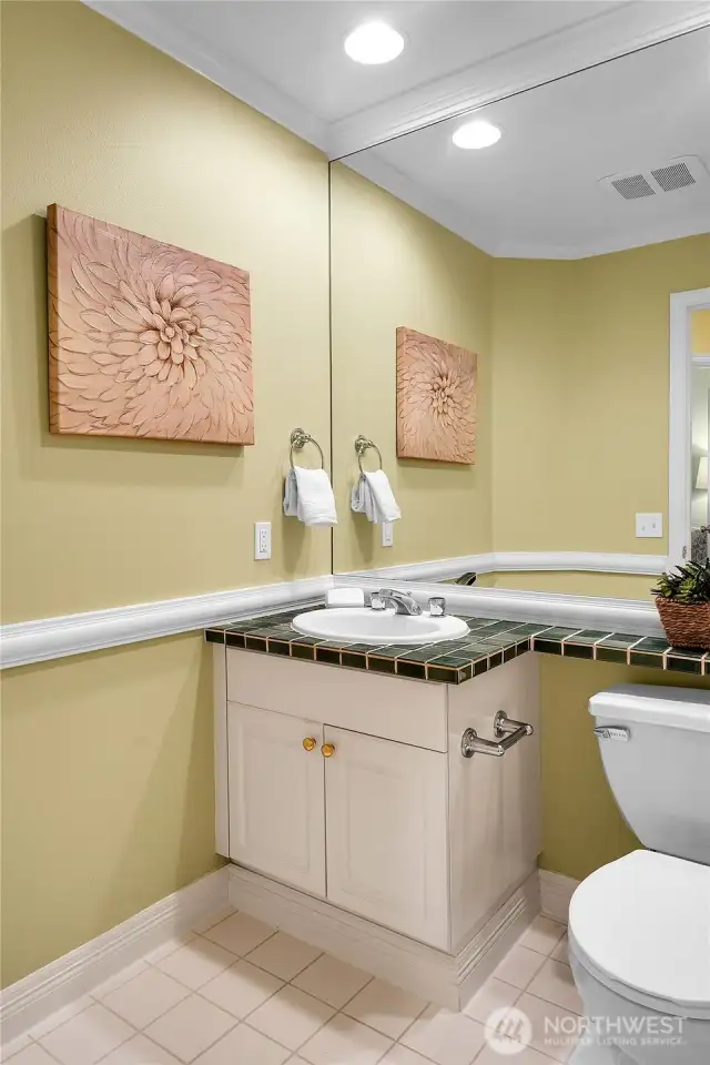 Powder bath off entry hallway. Gerber®-brand watercloset with banjo-style tiled countertop. Paneled white cabinets with shiny-brass hardware, wall-to-wall vanity mirror above. Crown molding. Oak-hardwood floor.