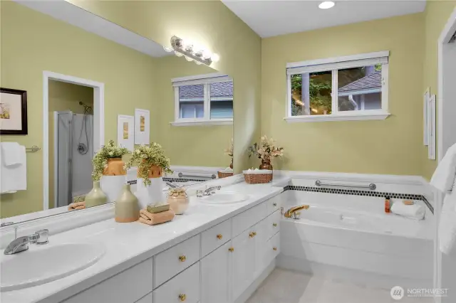 Primary bath off bedroom. Banjo-style tiled countertop at double sink vanity. Tile floor. Paneled white cabinets with shiny brass hardward, brass-accented shiny-chrome faucets. Shiny chrome style light bar with globe lights. Vinyl window with window screen and two-inch blinds. Oval tub with safety hand bar, tile surround and Traditional millwork. Paneled Quaker pocket door to watercloset room with tile floor. American Standard® elongated watercloset. Fiberglass-molded shower with clear glass doors and handheld shower wand. Large walk-in closet with coated ventilated wire organizer shelving.