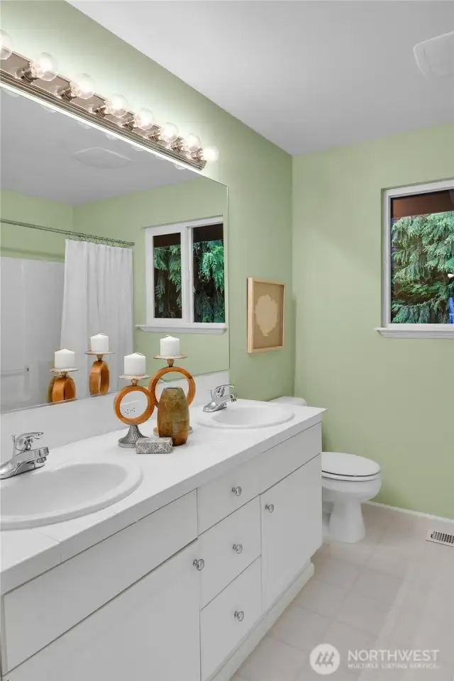 Upper-level guest bathroom. Paneled Quaker door. Tiled double-sink vanity with shiny chrome fixtures, paneled white cabinet doors, shiny-chrome hardware. Mansfield® watercloset. South-facing window with window screen. Fiberglass bathtub with safety hand bar, and shower. Linen closet with coated ventilated wire shelving. Vinyl floor.