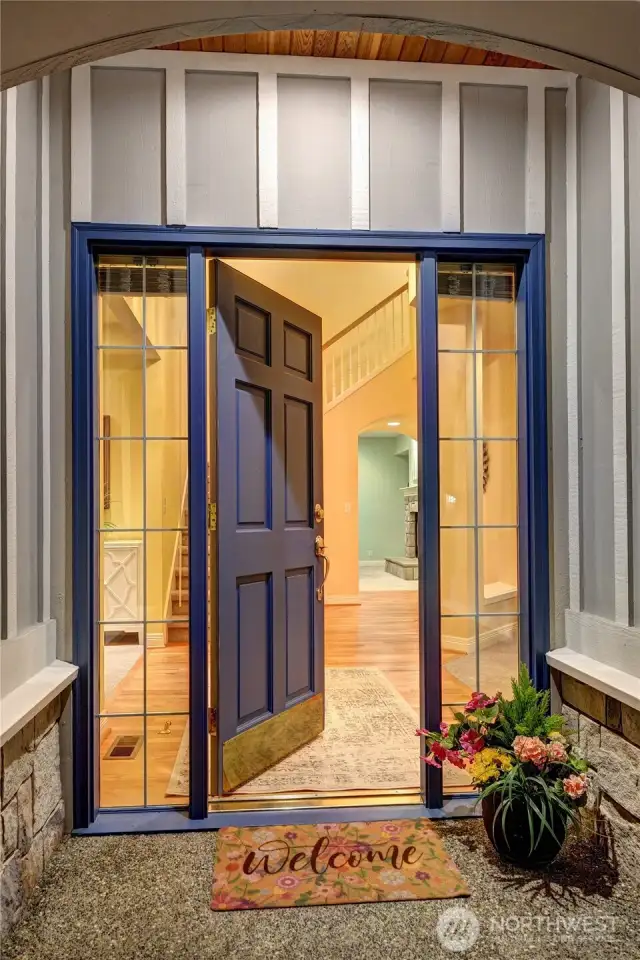 Entry exterior. Aggregate walkway and one slight step to the covered front porch with Tudor-style entry-door archway. Paneled Quaker door with brass kick plate, brass thumb-latch door hardware, deadbolt lock. Divided-light sidelight windows with blinds.
