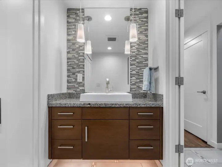 The remodeled powder room exudes refined elegance, featuring a floating vanity with vessel sink, granite countertops, a mosaic tile backsplash, and designer pendant lighting, with a discreetly integrated washer and dryer closet for added convenience.