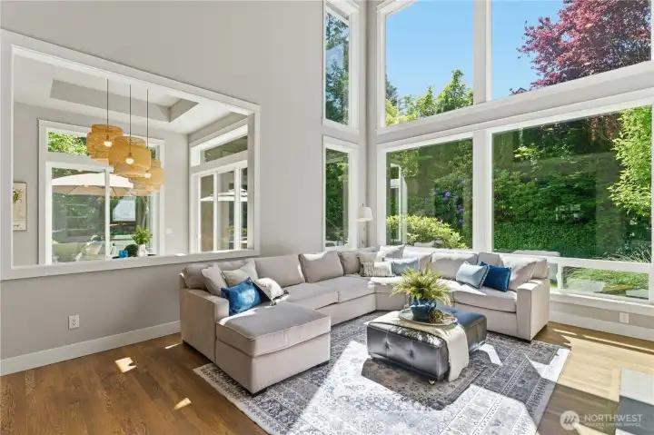 A bright, open gathering space with garden views, rich hardwood floors, and a soaring two-story ceiling that adds volume and light