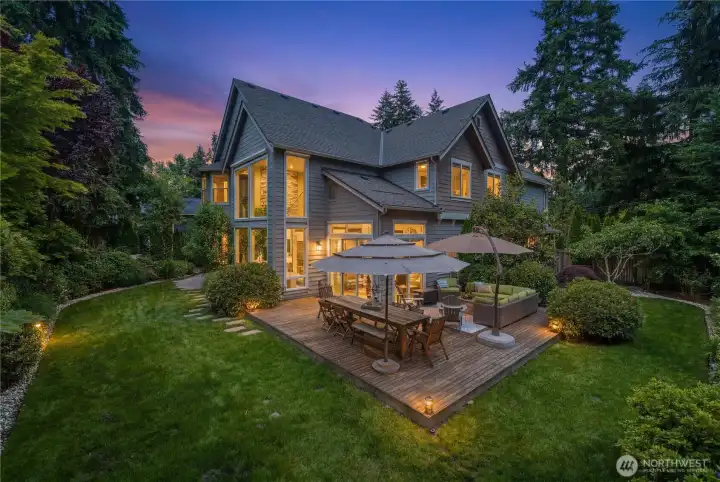 As dusk settles, the cedar deck and lawn glow with warmth. The patio rests privately to the right among the trees, hidden from this vantage point