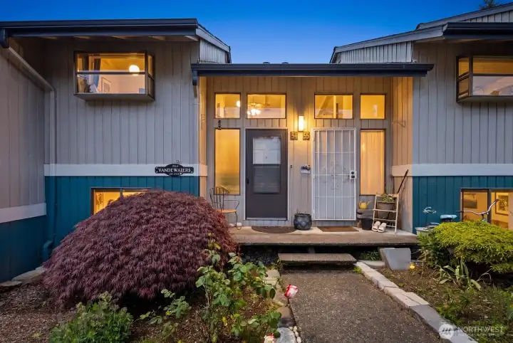 Welcoming townhouse-style entry with landscaped garden beds, covered front porch, and warm evening lighting.