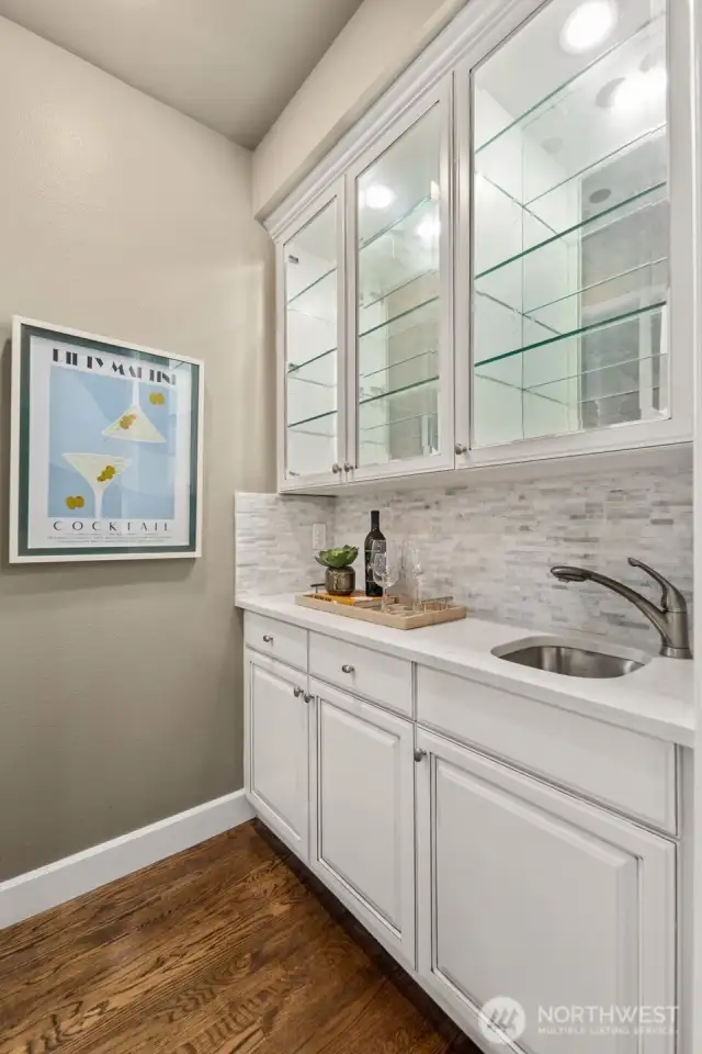 Stylish wet bar with glass-front cabinets and marble backsplash, seamlessly connecting the kitchen and dining room-perfect for entertaining.