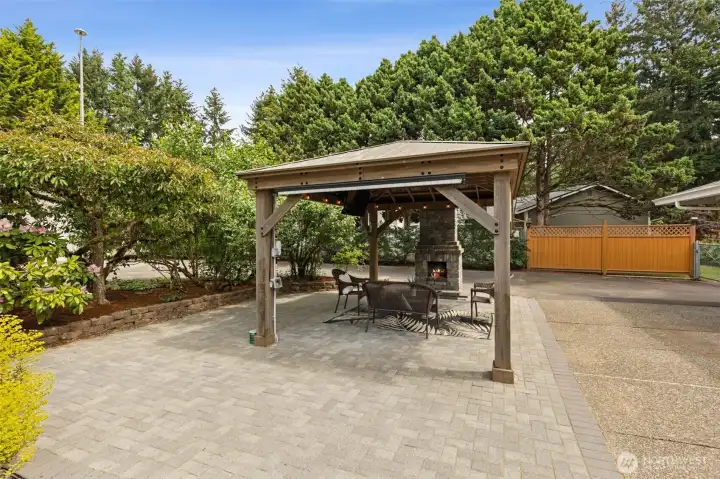 12x16 gazebo featuring a mounted TV, string lights, built-in wood-burning fireplace, and sunscreen shades for year-round enjoyment.