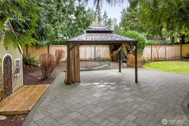 Covered gazebo lounge anchors the paver patio, offering a comfortable all season hangout space surrounded by greenery and garden beds.