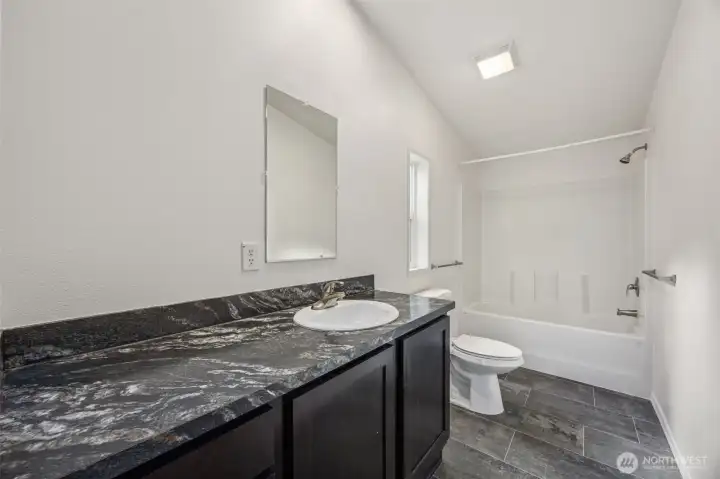 This en suite bathroom understands balance. The long vanity offers real counter space, paired with a dark shaker cabinet that feels modern and substantial. The marble-look Formica countertop is durable, forgiving, and refreshingly low stress. Matching brushed nickel hardware ties the room together, while the privacy-glass window adds daylight and airflow without sacrificing discretion. Stone-style vinyl flooring finishes the space with durability and polish.