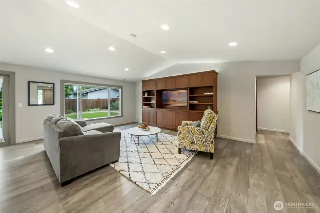 Notice the vaulted ceilings that add depth and openness to the space, complemented by a large picture window with automated blinds for light control and privacy. You'll find the primary suite, utility room, two bedrooms, and the main bathroom down the hall you see in the right of this photo.