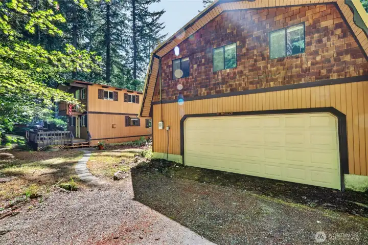 Alternate front elevation highlighting the attached garage, paved driveway, and mature evergreen surroundings. Alternate front elevation highlighting the attached garage, paved driveway, and mature evergreen surroundings.