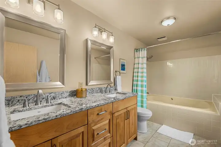Updated primary bath featuring dual vanities with granite countertops, warm wood cabinetry, and modern fixtures