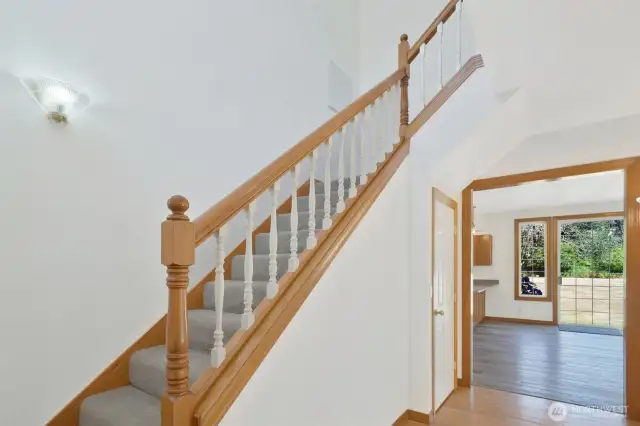 Classic stairway with warm wood tones and white spindles. View extends into the open-concept kitchen with large windows and outdoor access.