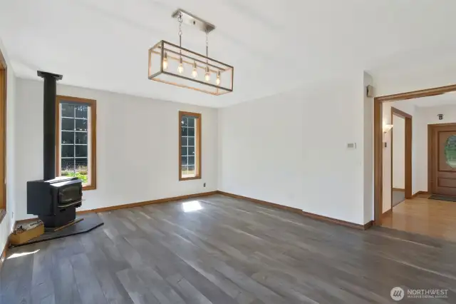 Bright and open living room featuring luxury plank flooring, modern light fixture, and a cozy wood-burning stove nestled between tall windows for scenic outdoor views.