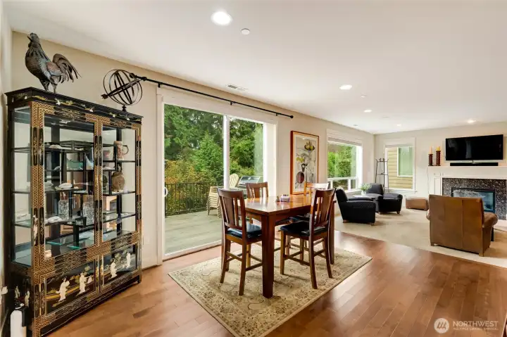 The open dining and living area flows effortlessly to the outdoor deck through large sliding doors, creating a seamless indoor-outdoor living experience. Natural light fills the space, highlighting the beautiful hardwood floors and peaceful, tree-lined backdrop.