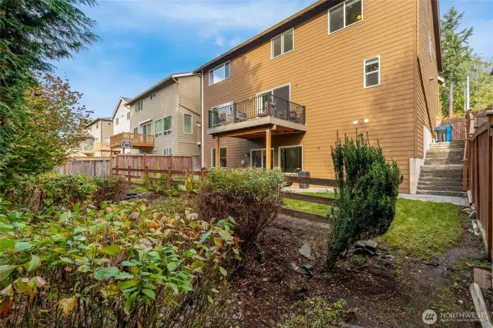 Rear elevation view highlighting the covered patio, upper deck, and fully fenced yard surrounded by mature landscaping for privacy.