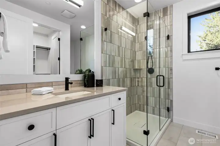 Upper level ensuite bathroom. Large vanity with slab counters, framed mirror, shower with frameless glass shower doors, a shampoo shelf, and a walk-in closet.