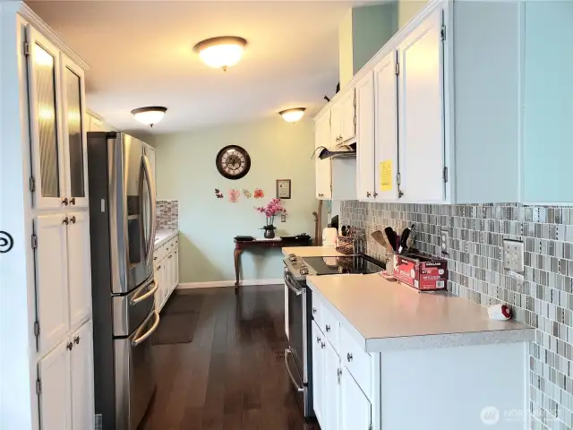 Opposite View of the Huge Walk Through Kitchen with Tile Back Splash, White Cabinets, Stainless Appliances, Newer Paint and  Hard Wood Flooring Looking at the Eating Area.