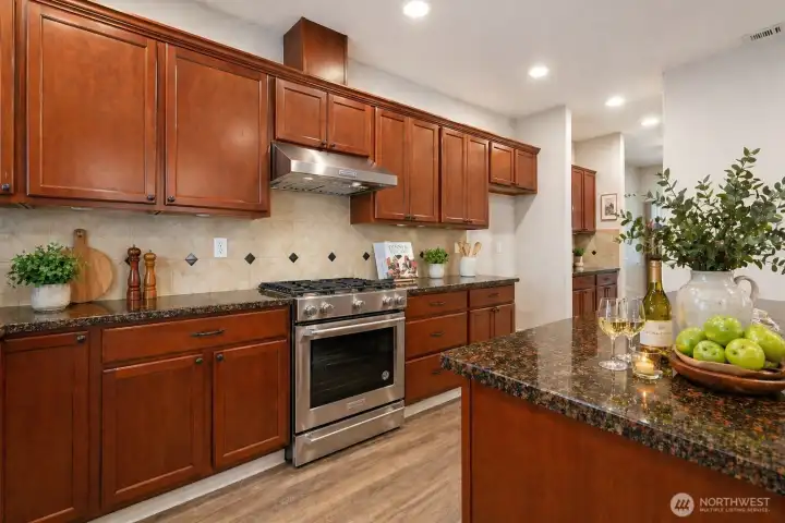A chef-inspired kitchen showcasing rich wood cabinetry, gleaming granite countertops, a stylish tile backsplash and large planks laminate flooring. Featuring a stainless-steel gas range with hood, a large island, and abundant storage, this space is both beautiful and highly functional. Open to the dining and living areas with warm laminate floors and recessed lighting, it’s perfectly designed for everyday living and effortless entertaining.