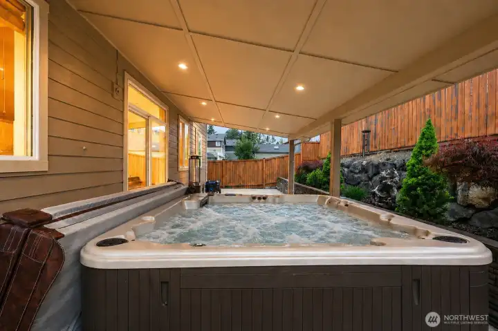 Unwind in your own private backyard retreat featuring a luxurious covered hot tub spa, perfectly designed for year-round relaxation. Surrounded by warm ambient lighting, privacy fencing, and beautifully landscaped retaining walls, this inviting outdoor space creates a peaceful resort-style atmosphere ideal for quiet evenings or entertaining guests in comfort and style.