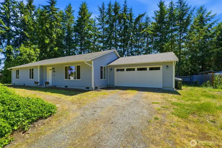 Classic ranch-style curb appeal with two-car garage, gravel driveway, and towering Pacific Northwest evergreens framing the home.