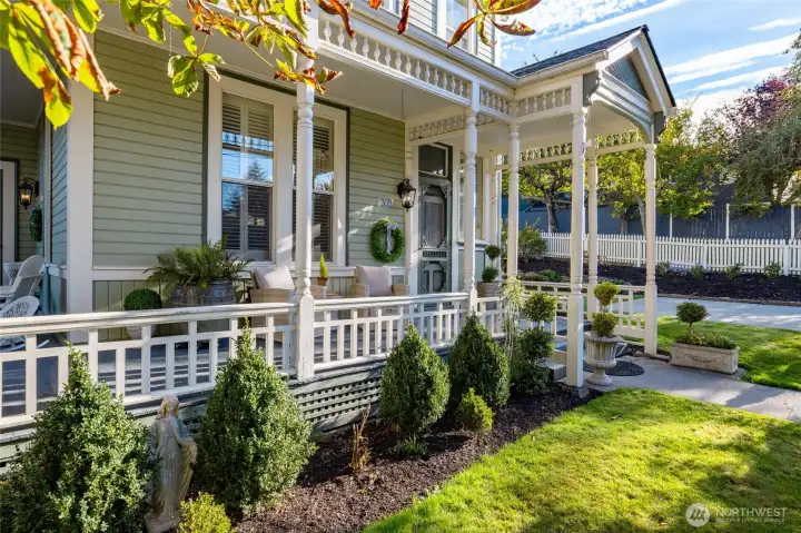 A charming, manicured front garden framed by a white picket fence, mature shade tree, and the home’s picturesque Victorian porch with detailed spindlework.