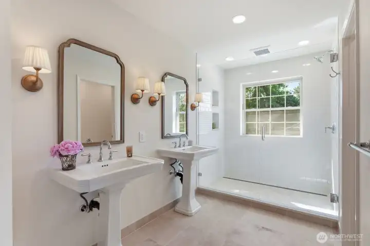 A brand new primary bathroom offering travertine flooring, oversized shower and dual pedestal sinks with brass sconce lighting.