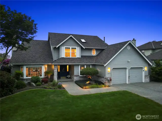 This beautifully maintained home features a refined gray exterior with crisp white trim and a stylish dark gray roof. A spacious double garage sits conveniently to the right, framed by a wide concrete driveway for ample parking. The home's inviting front entrance, accented by warm exterior lighting, opens to a carefully landscaped yard with lush greenery and neatly trimmed shrubs. Large windows add to its bright and welcoming aesthetic, while the evening glow creates a cozy and inviting ambiance.