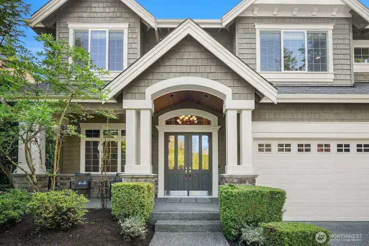 The grand arched entryway makes a striking statement with its elegant glass-paneled double doors, rich stone surround, and craftsman-inspired detailing. Lush flanking greenery and classic column accents create a warm and inviting welcome the moment you arrive.