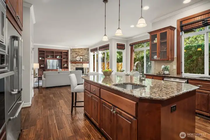 This beautiful perspective highlights the seamless open-concept flow between the gourmet kitchen and the great room beyond, showcasing the expansive island, gleaming granite countertops, and warm acacia hardwood floors that unite the spaces. Abundant natural light pours in through large windows, creating a bright and airy atmosphere perfect for both everyday living and effortless entertaining.