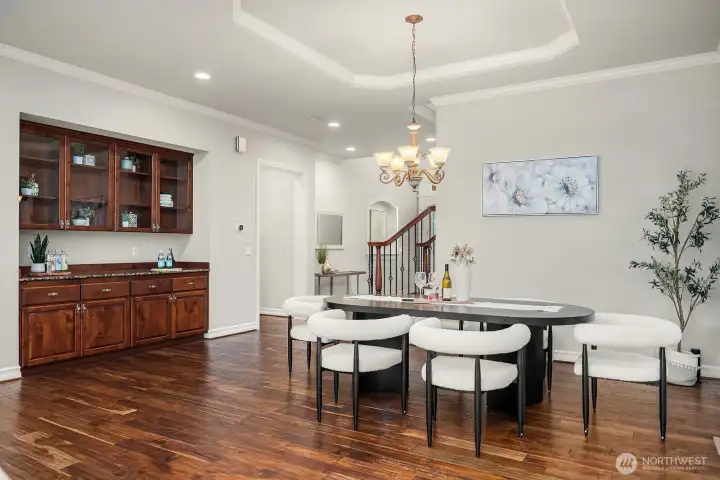 This angle of the formal dining room reveals a stunning built-in butler's pantry with rich cabinetry and glass-front display doors, adding both elegance and exceptional storage and serving convenience. The tray ceiling, statement chandelier, and gleaming hardwood floors continue to reinforce the refined, upscale character of this beautifully appointed home.