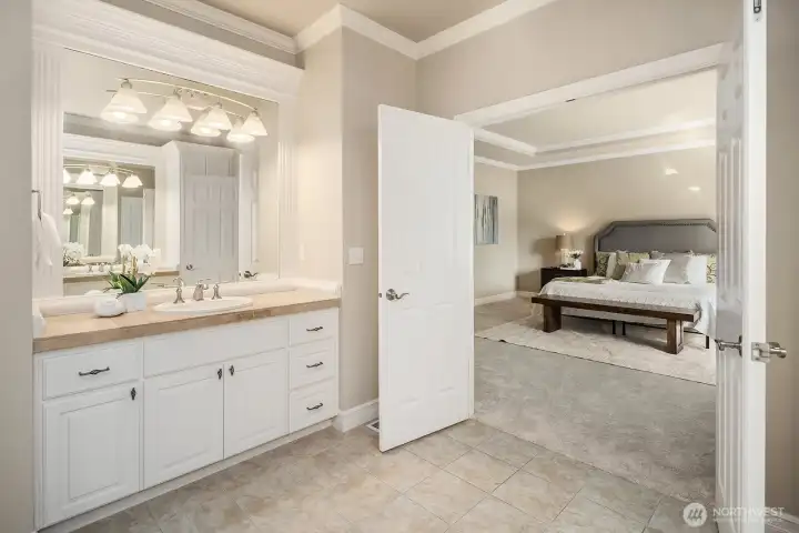 This view of the primary bath beautifully captures the seamless connection between the spa-inspired ensuite and the expansive primary suite beyond, highlighting the thoughtful and functional layout of this private retreat. The dual vanities with crisp white cabinetry, gleaming countertops, and elegant tray ceiling detail reflect the exceptional quality and craftsmanship found throughout the home.