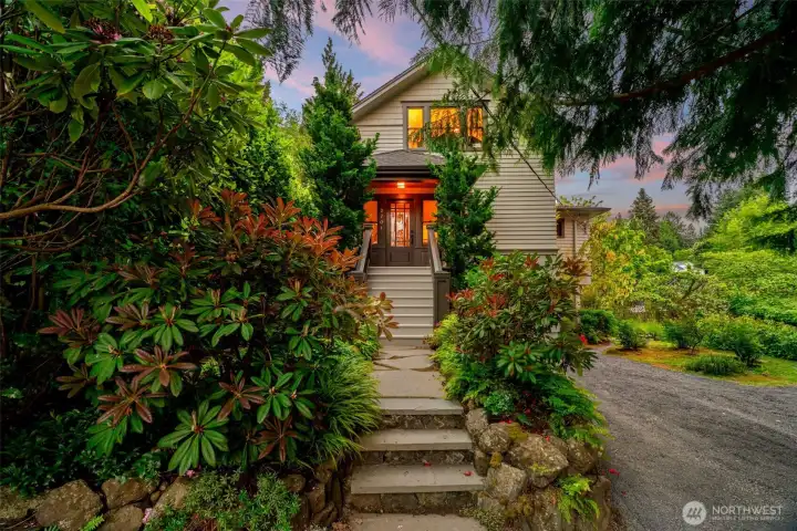First impressions don't get more enchanting than this — a warmly lit front entry framed by lush rhododendrons and towering evergreens welcomes you home under a painted evening sky.