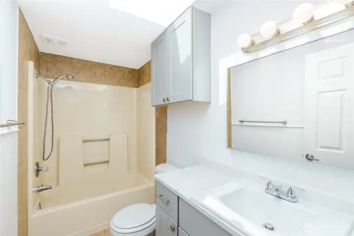 The updated guest bathroom features a modern vanity with a quartz-style countertop, fresh cabinetry, and a convenient tub/shower combination with built-in seating and grab bars, offering comfort, functionality, and a clean, contemporary finish.