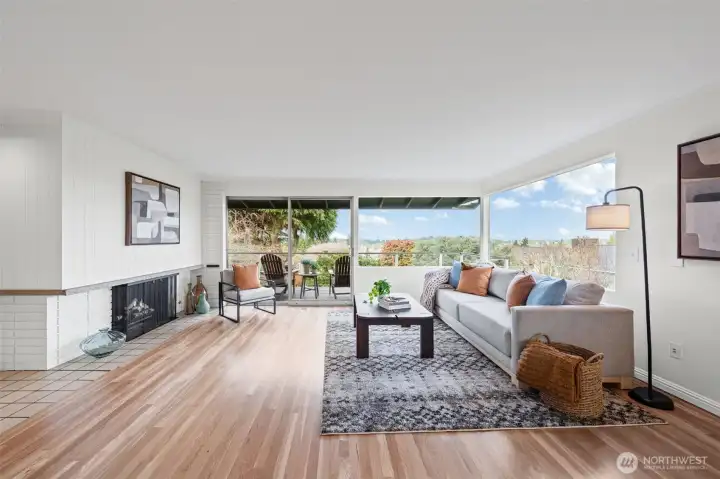 Light-filled living room with expansive windows and seamless indoor-outdoor flow to a private deck with territorial views. Warm hardwood floors and a cozy fireplace create an inviting space for everyday living and entertaining.