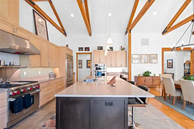 View 2 of the Living Room and Dining Room with soaring ceilings, exposed wood beams, wide plank wood floors, stone fireplace and glass sliding doors leading to the outdoor living area.