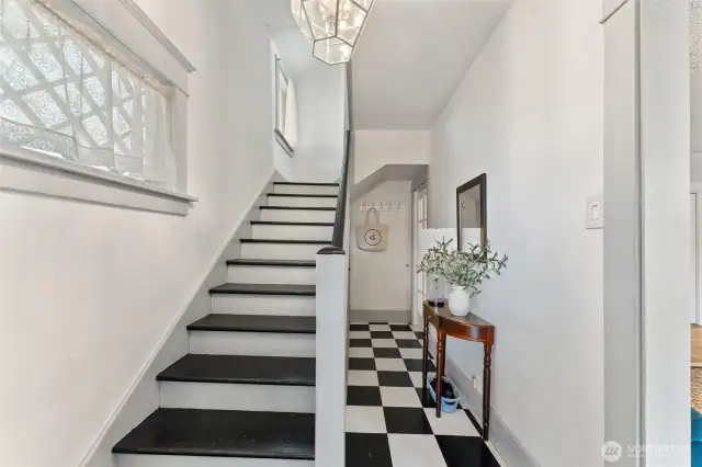 Welcoming entryway with black-and-white checkerboard flooring, bright natural light, and a classic staircase leading upstairs to two bedrooms, a bathroom and sitting area.
