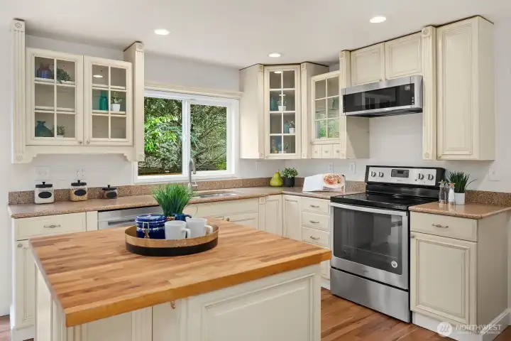 Charming kitchen detail highlighting the warm butcher block island, glass front cabinetry, stainless steel appliances, and lush greenery visible through the window. Charming kitchen detail highlighting the warm butcher block island, glass front cabinetry, stainless steel appliances, and lush greenery visible through the window.