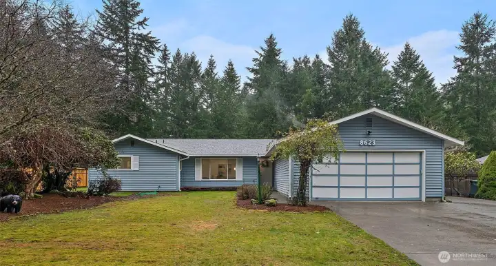 Charming ranch-style home with fresh blue-gray siding, attached two-car garage, and a lush evergreen backdrop that adds natural privacy and PNW character.