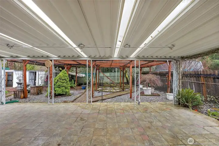 A large covered patio with a translucent roof and stone tile flooring extends the living space outdoors year-round, overlooking a mature garden with a wood pergola and established plantings.